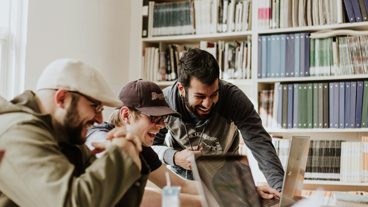 Three college-aged men gather around two laptops in a library.