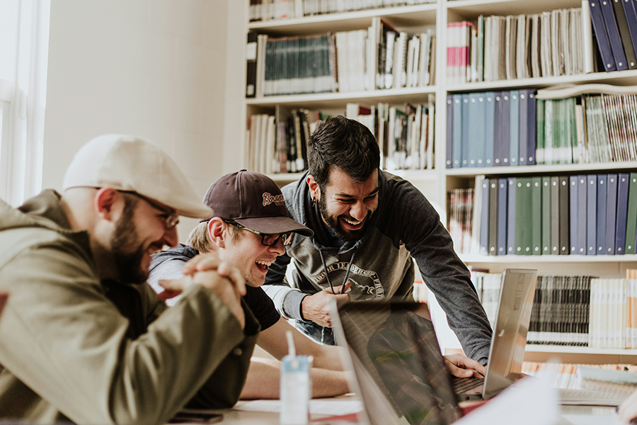 Three college-aged men gather around two laptops in a library.