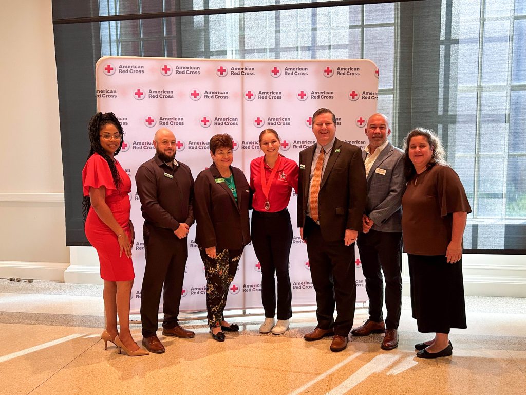 A group of 7 men and women stand for a picture in front of a white banner with the American Red Cross words and its logo of a white circle with a red plus sign inside.