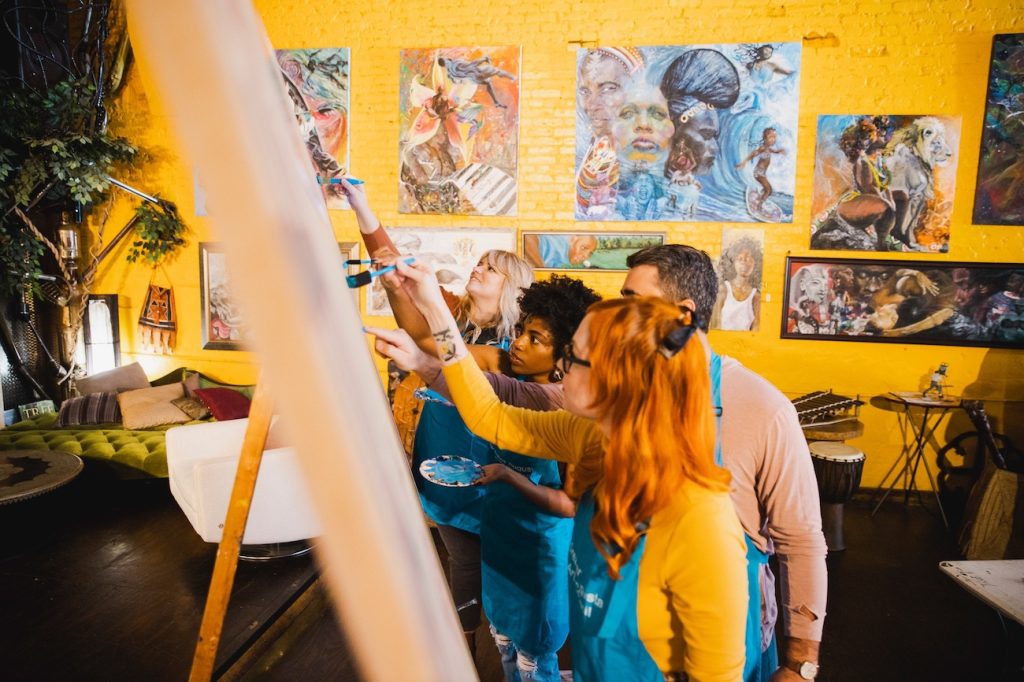 Four people paint canvases on easels in a room with golden yellow walls and paintings covering the walls..