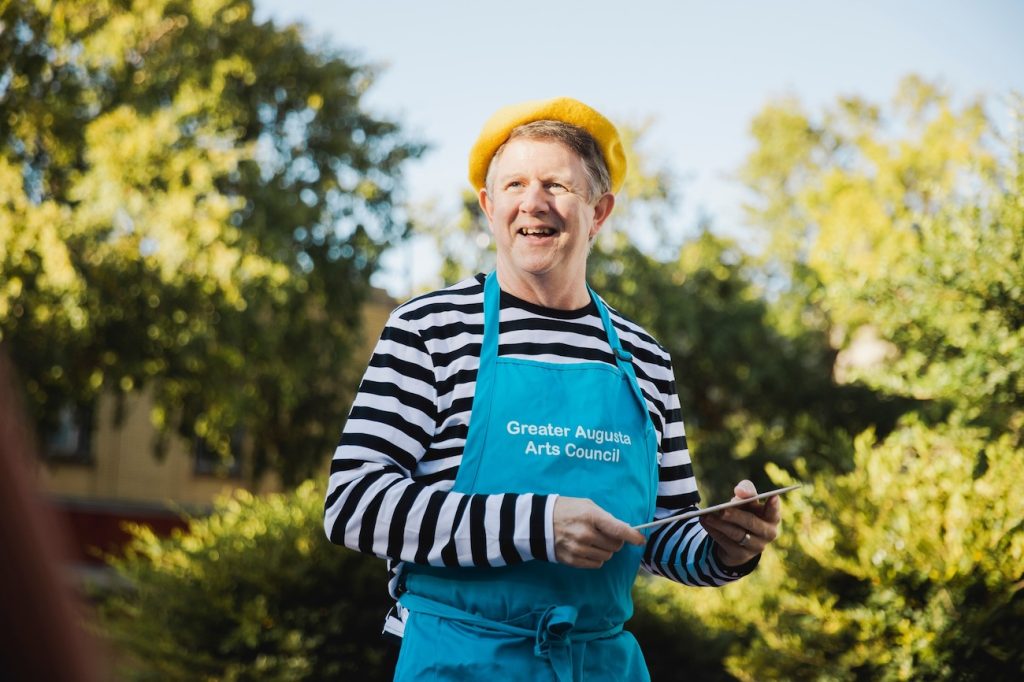 A man wearing a yellow beret, black and white striped shirt and blue apron with the words "Greater Augusta Arts Council" in white lettering holds a palette outside while smiling at something off camera.