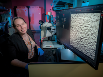 A woman sits at a computer with a microscope next to her.