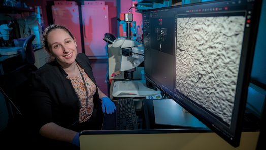 A woman sits at a computer with a microscope next to her.