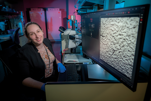 A woman sits at a computer with a microscope next to her.