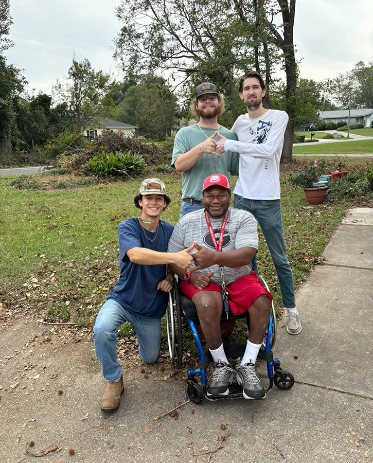 Three male college students pose for a photo with a wheelchair-bound homeowner after they cleared his yard of debris after a natural disaster.