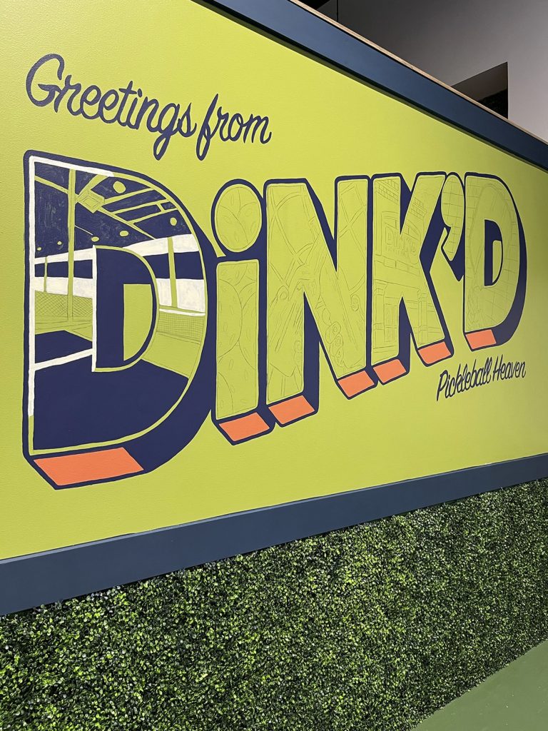 A progression of a mural in a pickleball facility with the words "Greetings from DiNK'D Pickleball Heaven." The "D" is now filled in.