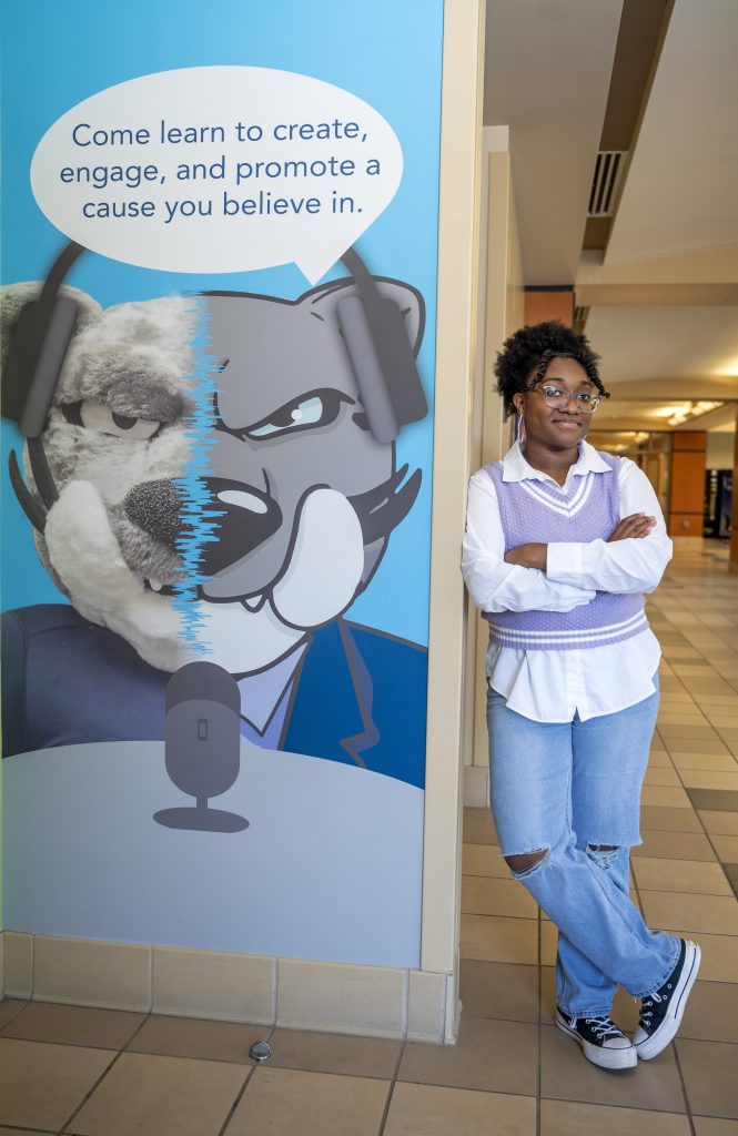 A college woman leans against a wall featuring a mural she created. The mural features a college mascot in the form of a jaguar.