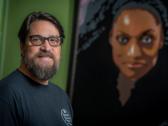 A man wearing glasses stands in front of a mural of a woman.