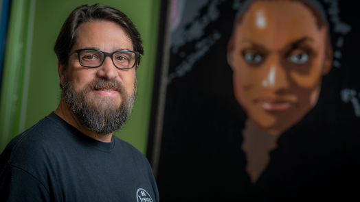 A man wearing glasses stands in front of a mural of a woman.