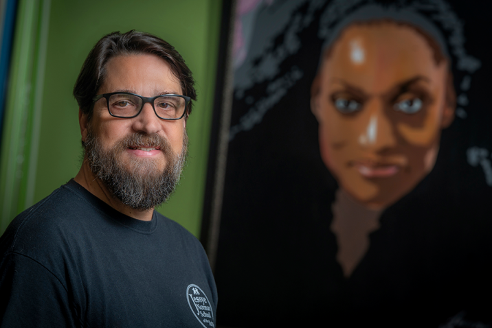 A man wearing glasses stands in front of a mural of a woman.