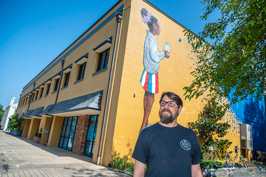 A man wearing glasses stands outside in front of a building that is painted with a mural of a young girl holding a glass jar as butterflies fly away.