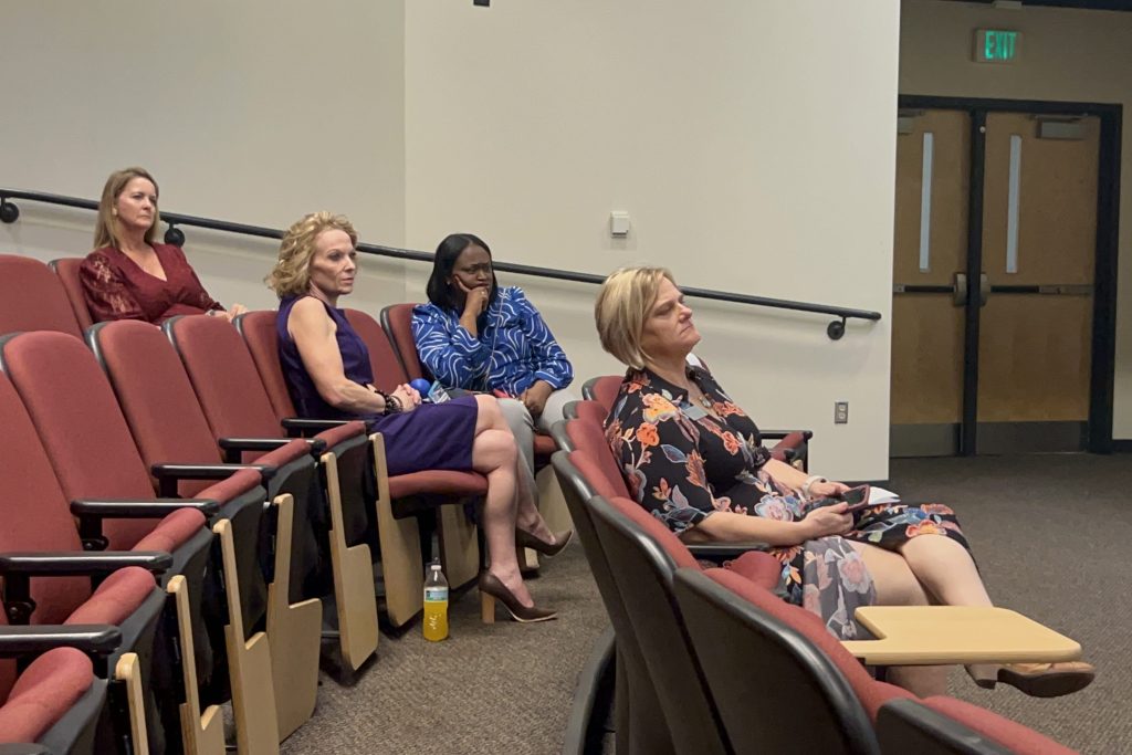 Four women sit in a large auditorium listening to a speaker.