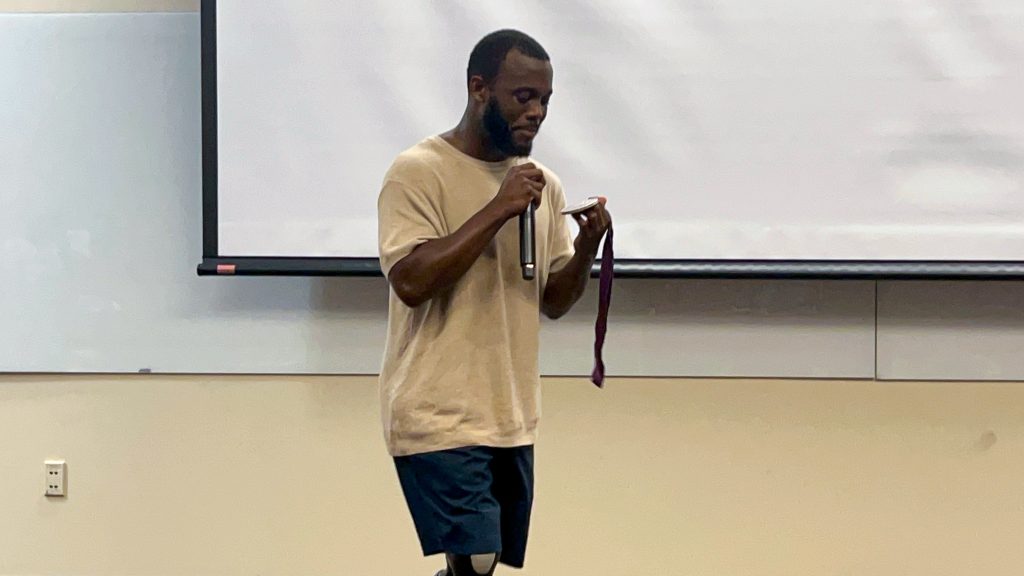 A man is holding a microphone and an Olympic medal while speaking in an auditorium.