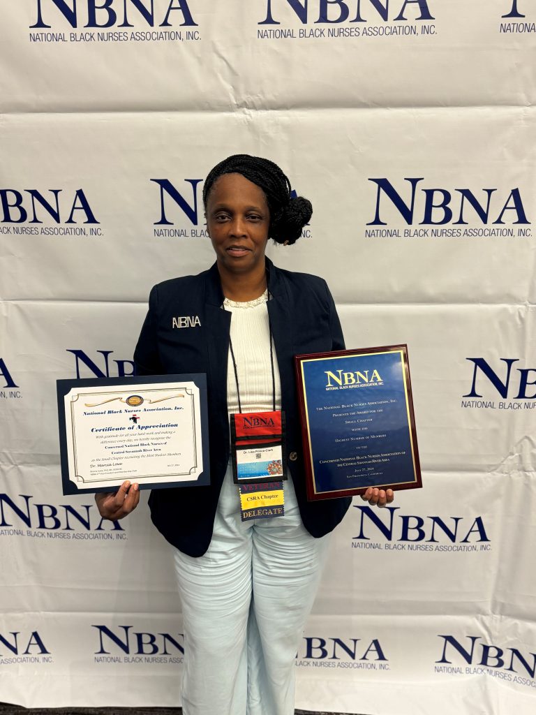 A woman wearing a black jacket with the white initials NBNA, a white shirt and light blue pants hold an award in each hand while standing in front of a white backdrop with the words NBNA National Black Nurses Association"