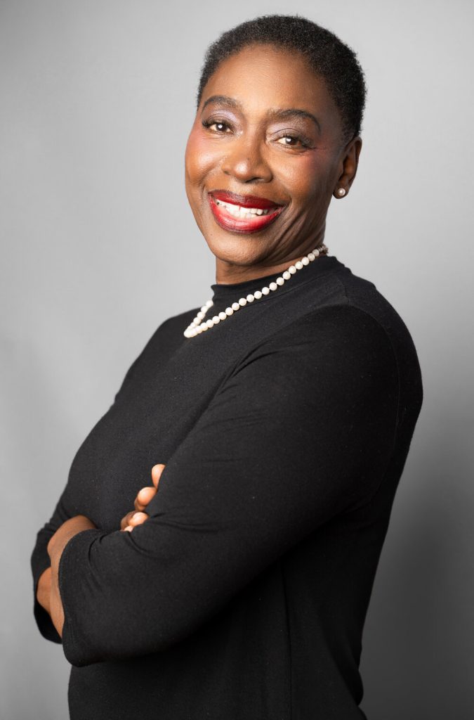 A woman dressed in all black with a white pearl necklace crosses her arms and smiles for the camera.