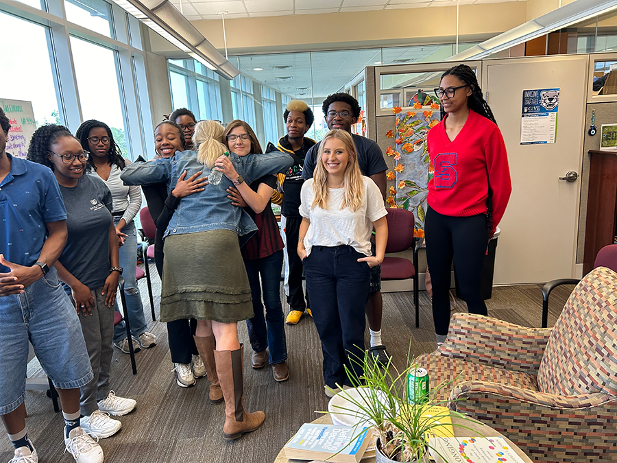 A group of college students gather in an office area while a woman in the front hugs the students in the middle.