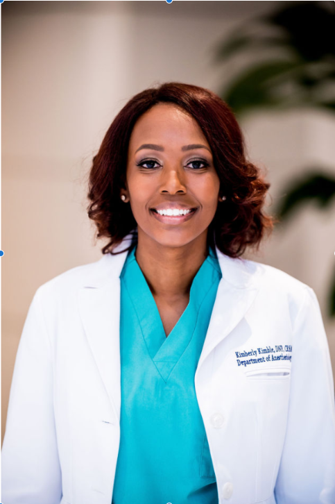 A woman wearing a teal shirt and white coat with her name, credentials and department name smiles at the camera