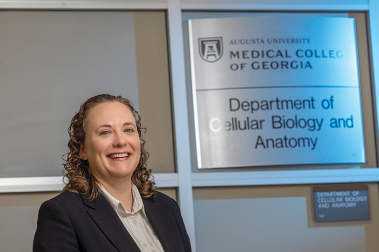 A woman stands next to a plaque that reads Augusta University Medical College of Georgia Department of Cellular Biology and Anatomy