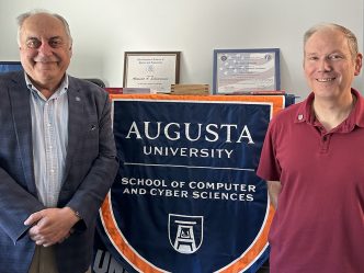 Two men stand near a flag that says "Augusta University School of Computer and Cyber Sciences."