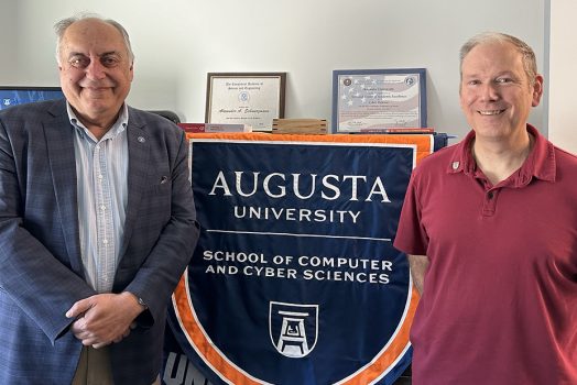 Two men stand near a flag that says "Augusta University School of Computer and Cyber Sciences."