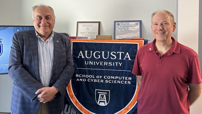 Two men stand near a flag that says "Augusta University School of Computer and Cyber Sciences."