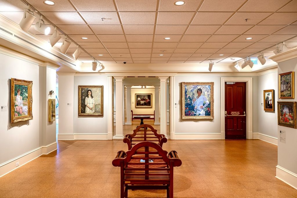 Chairs sit in the center of a large room with various painting hanging on the walls around the room of a museum.