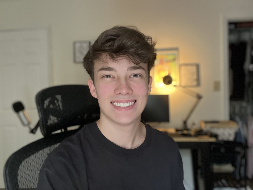 A male sitting in a computer chair in a room smiles at a camera