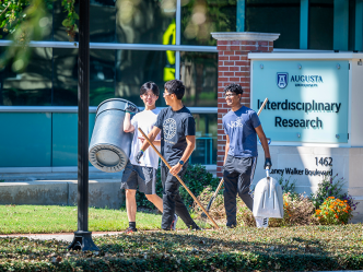 Three male college students walk outside with a large trash can and gardening tools while helping clean up a college campus.