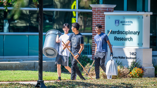 Three male college students walk outside with a large trash can and gardening tools while helping clean up a college campus.