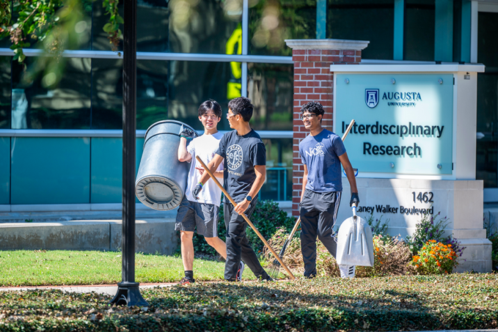 Three male college students walk outside with a large trash can and gardening tools while helping clean up a college campus.