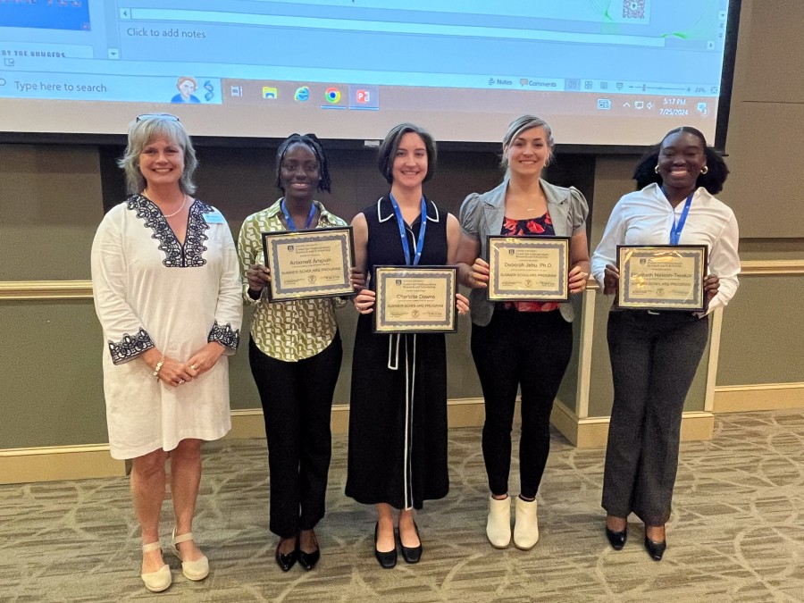 A woman stands with four female students holding awards at the front of a large room.