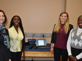 Three female students and a female professor stand in front of a table smiling at the camera.