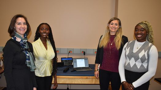 Three female students and a female professor stand in front of a table smiling at the camera.