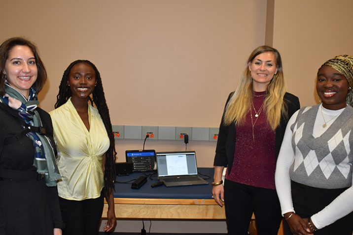 Three female students and a female professor stand in front of a table smiling at the camera.