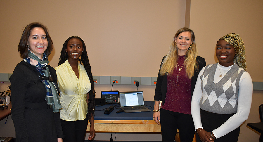 Three female students and a female professor stand in front of a table smiling at the camera.