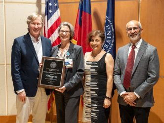 Four people pose for a photo together while a woman in the middle holds up an award plaque.