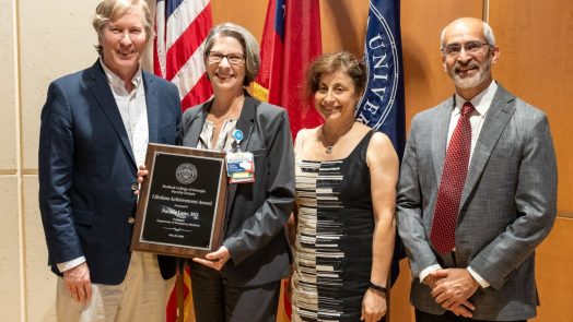 Four people pose for a photo together while a woman in the middle holds up an award plaque.