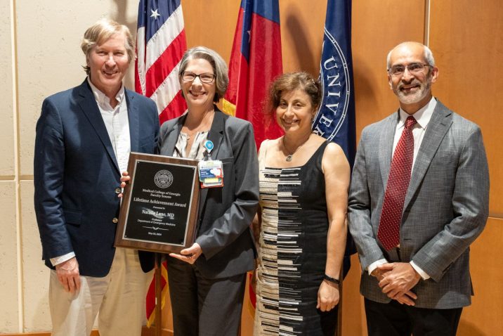 Four people pose for a photo together while a woman in the middle holds up an award plaque.