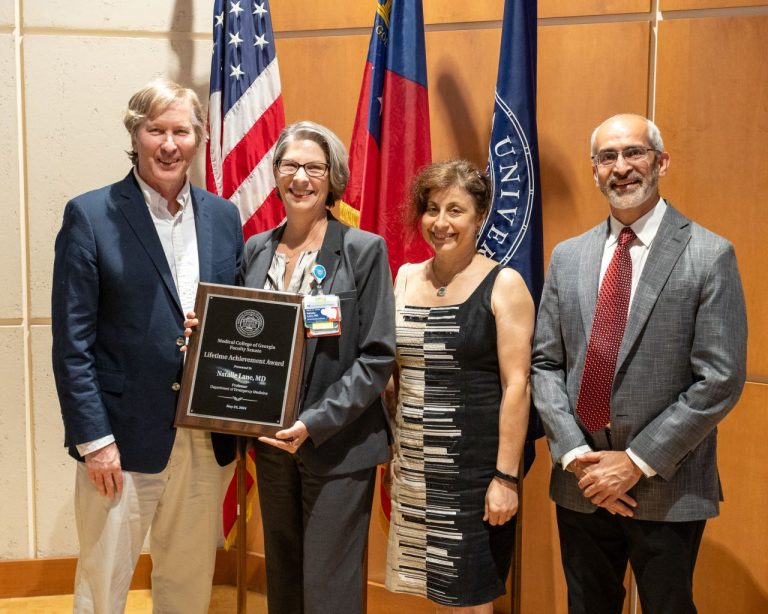 Four people pose for a photo together while a woman in the middle holds up an award plaque.