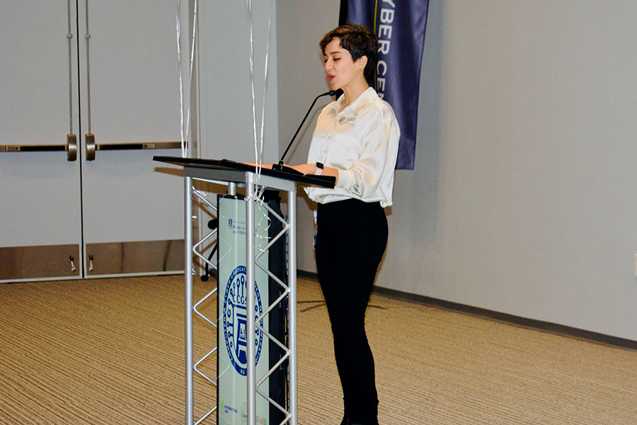 A woman stands at a podium with a microphone at the front of a large auditorium and speaks to a group of people.