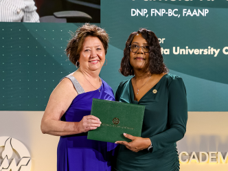 Two women stand on a stage. The woman on the right is handing the woman on the left an award.