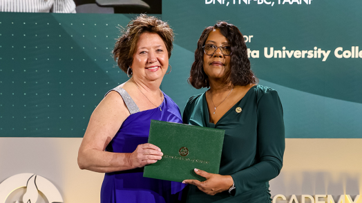 Two women stand on a stage. The woman on the right is handing the woman on the left an award.