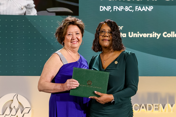 Two women stand on a stage. The woman on the right is handing the woman on the left an award.