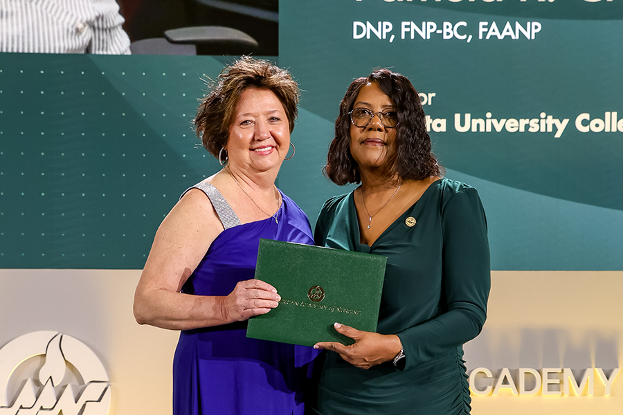 Two women stand on a stage. The woman on the right is handing the woman on the left an award.