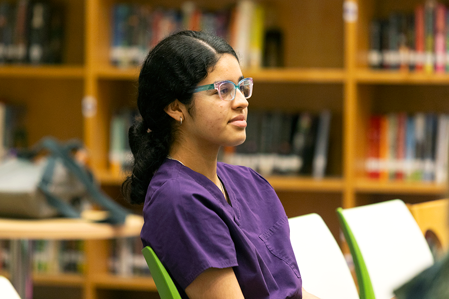 A high school girl wearing medical scrubs sits in a row of chairs and listens to a presentation.