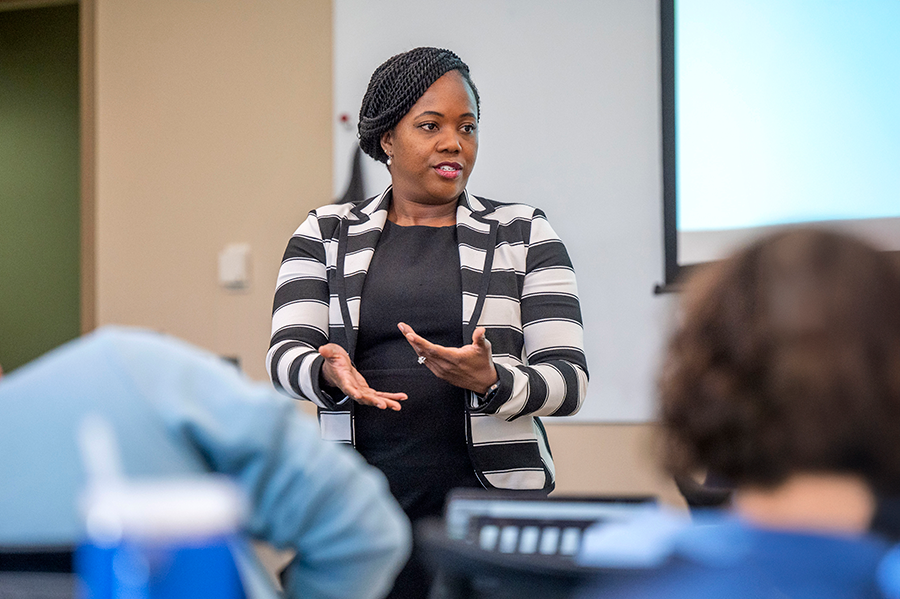 A female college professor stands at the front of a large college classroom an delivers a lecture to a group of students.