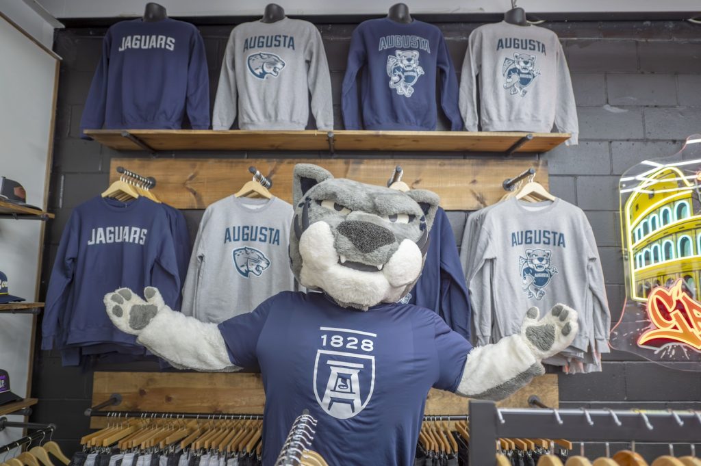 A college mascot depicting a large cat stands inside a small business selling clothing. There are several rows of shirts behind the mascot with that same mascot on them under the word "Augusta."