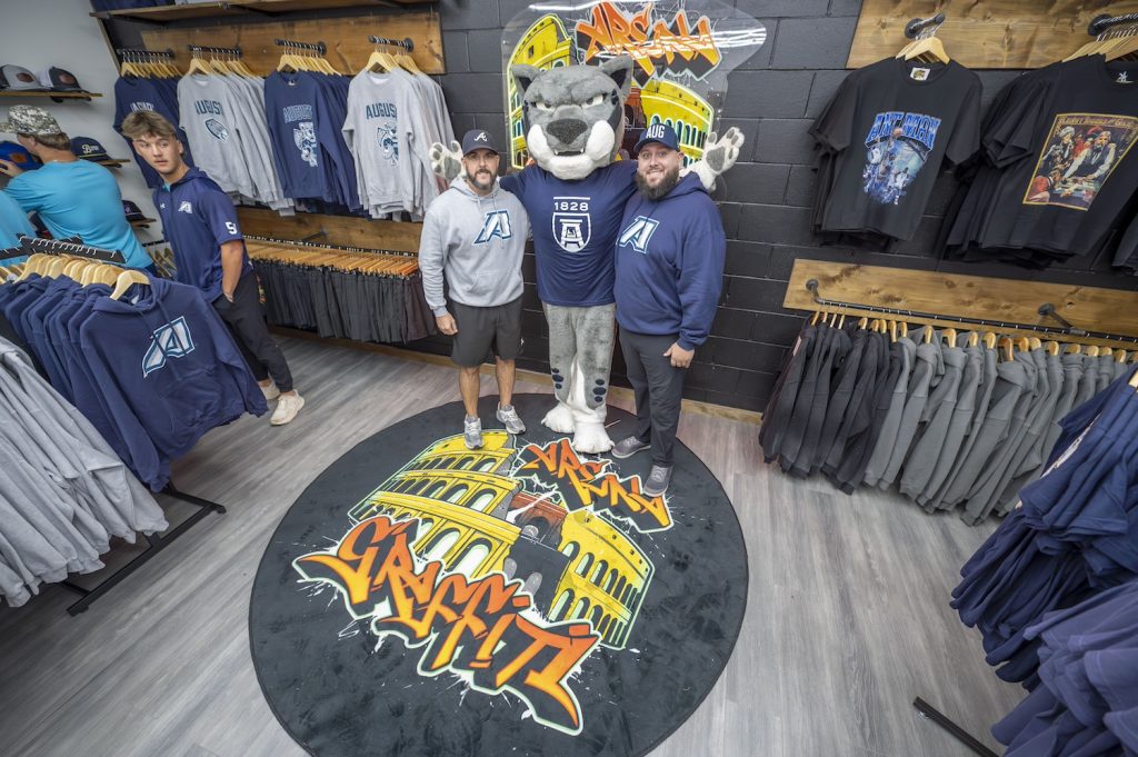 A college mascot depicting a large cat stands with two men inside a small business selling clothing.