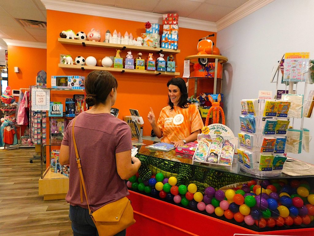 A female small business owner behind a check-out counter interacts with a customer inside a toy store.