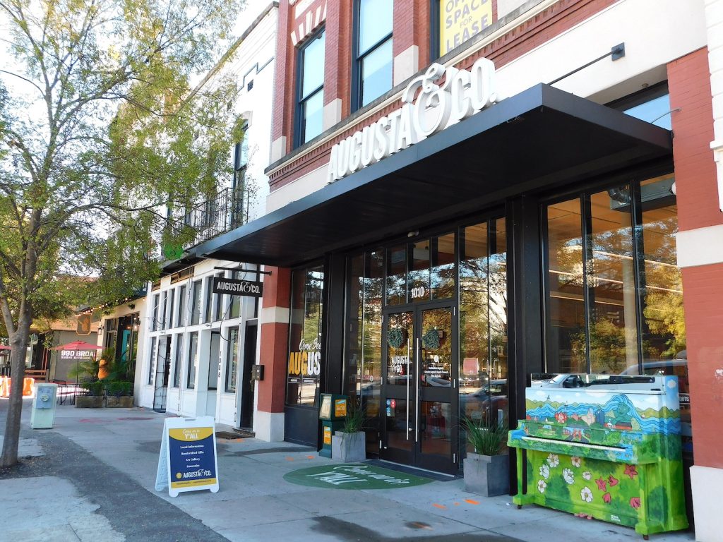 Storefront of a small business. There is a sign that reads "Augusta & Co." and a piano out front painted with flowers, a river and a small town.